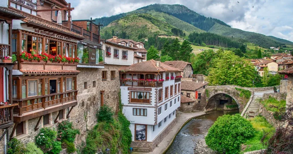 A charming village scene in Northern Spain, featuring stone buildings with wooden balconies adorned with flowers, a stone bridge over a river, and lush green hills, highlighting the beauty of Spain and Portugal's traditional architecture.