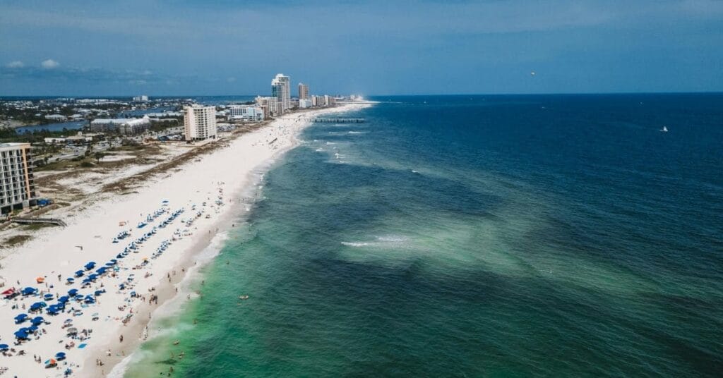 A scenic aerial view showcasing the things to do in orange beach alabama, including a pristine white sandy beach lined with colorful umbrellas, high-rise buildings, and the vibrant blue waters of the Gulf of Mexico.