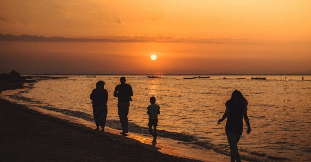 A serene sunset at Orange Beach, Alabama, with silhouetted figures walking along the shoreline and boats drifting in the distance.