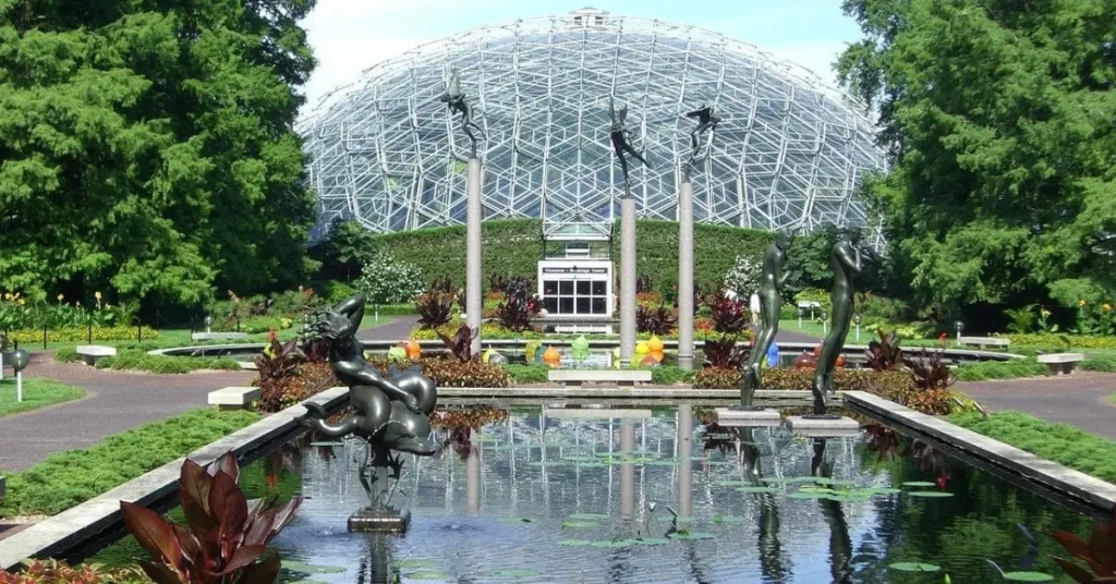 The Climatron at the Missouri Botanical Garden surrounded by lush greenery, sculptures, and a reflective pond.