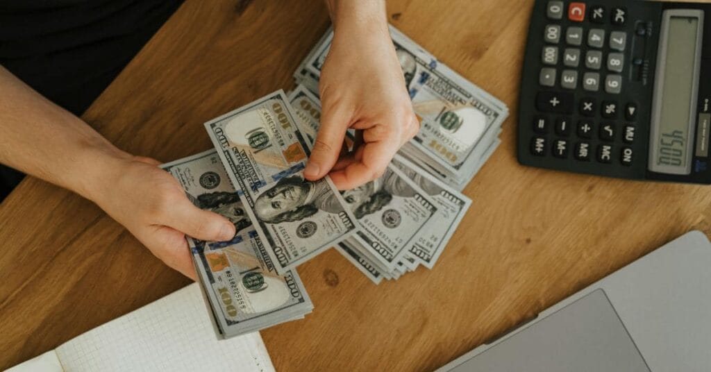 Hands counting money on a desk with a calculator and laptop, illustrating the financial aspect of becoming a travel agent.