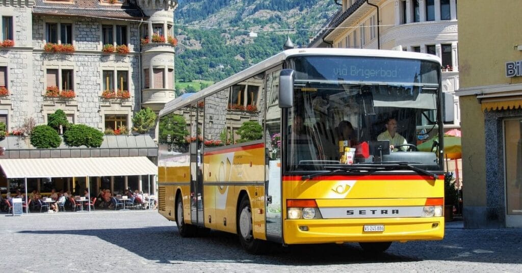 A bright yellow Setra bus traveling through a European town with cobblestone streets, historic buildings, and a scenic mountain backdrop.