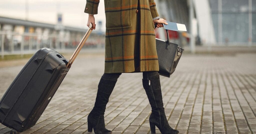 A woman in a stylish coat and boots carrying a black travel bag and pulling a suitcase.