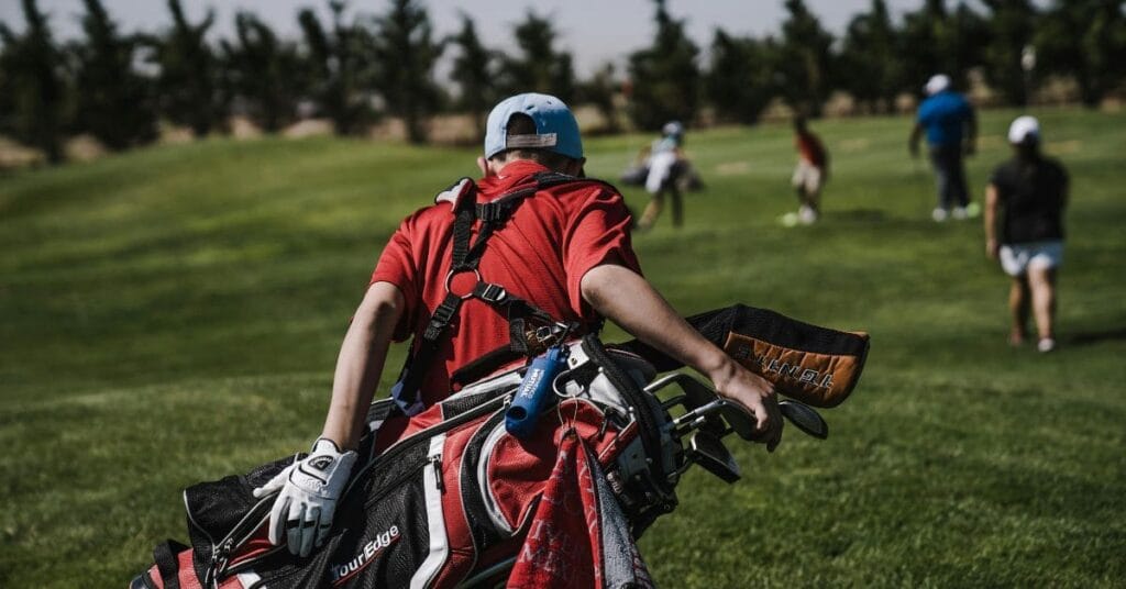 A young golfer carrying a golf bag across a lush golf course, heading toward other players on the green.