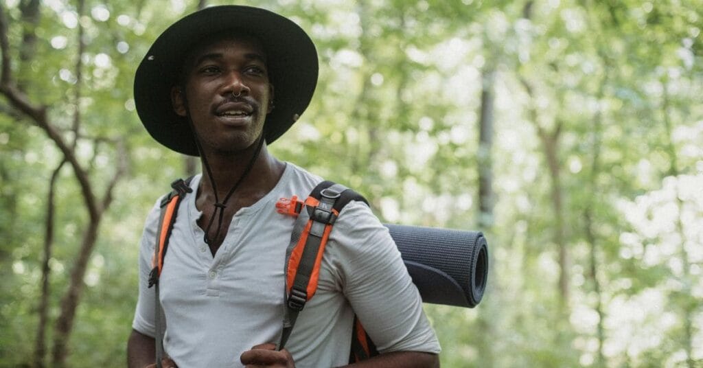 A single Black man wearing a wide-brimmed hat and carrying a backpack with a rolled-up mat, enjoying a hike in a lush green forest.