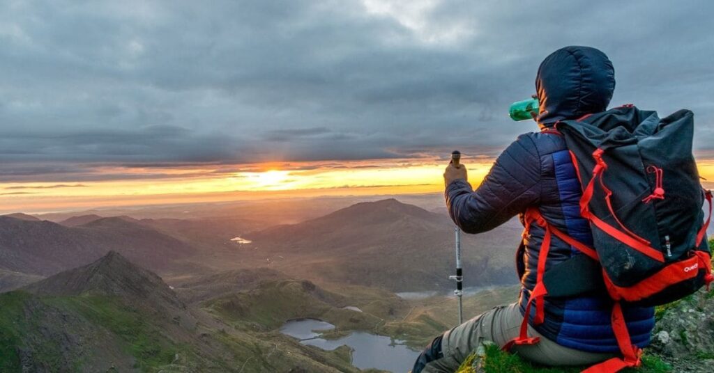 A single Black male traveler sitting atop a mountain peak, dressed warmly with a backpack and hiking gear, enjoying a serene sunset view, highlighting the best place for a solo Black man to travel.