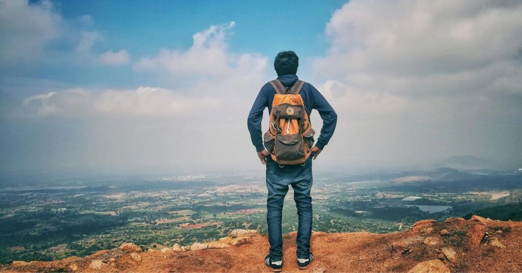 A single male traveler standing on a rocky cliff, wearing a backpack and gazing at a scenic view of fields and hills under a cloudy sky, symbolizing exploration and adventure, perfect for the best place for single black male to travel.