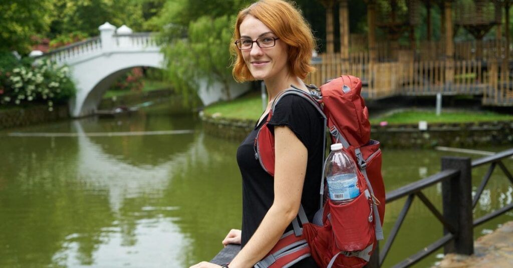 A smiling woman with a red backpack standing by a scenic pond with a white bridge in the background.