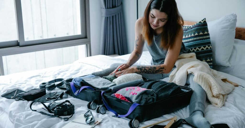 A young woman packing a suitcase on a bed, surrounded by travel essentials like clothes, a camera, and accessories.