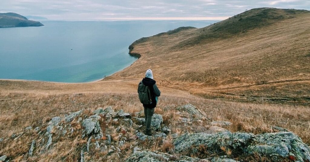 A solo traveler with a backpack standing on a rocky hill overlooking a serene lake and rolling grassy hills under an overcast sky, capturing the essence of nature and tranquility.