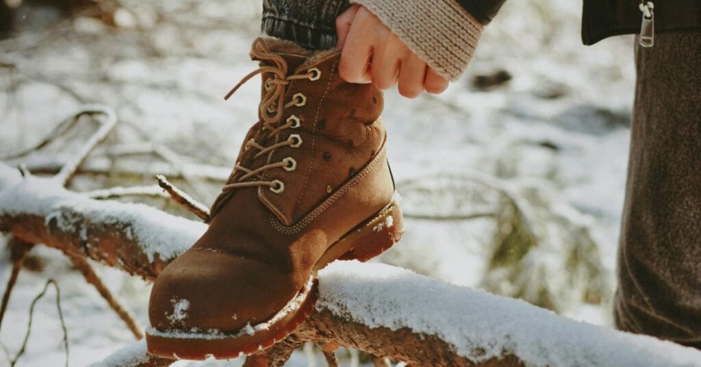 Person tying a brown winter boot on a snowy log, ideal for wide-foot comfort during winter travel, showcasing the best winter travel shoes wide feet will love.