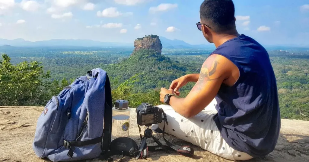 A traveler sitting on a hilltop with a backpack, camera gear, and water bottle, enjoying the scenic view of Sigiriya Rock in Sri Lanka.