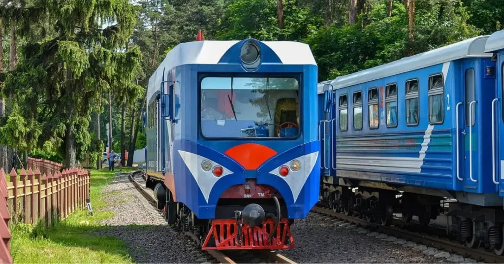 A train traveling from Aytown to Beetown, painted in vibrant blue and white, passing through a scenic green landscape with trees and a wooden fence alongside the railway tracks.