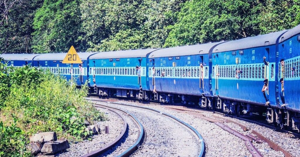 A long blue passenger train traveling from Aytown to Beetown, curving along railway tracks surrounded by lush greenery.