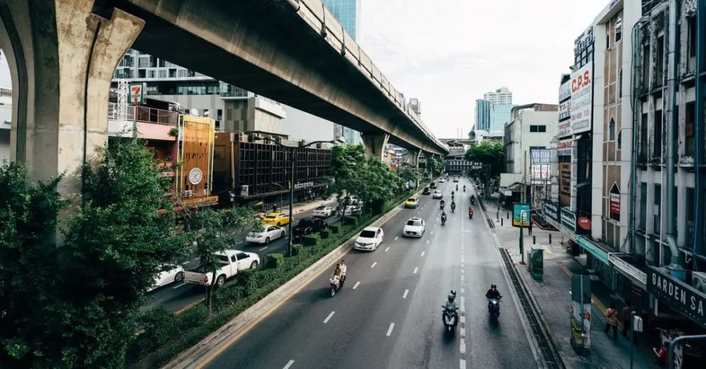 A bustling street in Bangkok, Thailand, with motorbikes, cars, and an elevated Skytrain track, highlighting the vibrant city life during the best time to visit Thailand.