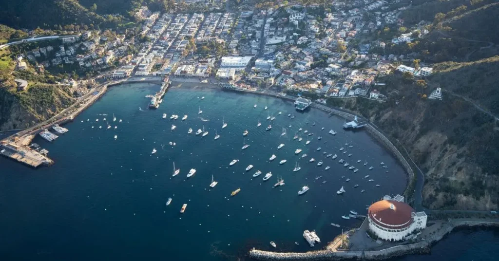 Aerial view of Avalon on Catalina Island, showcasing the Catalina Casino, a bustling harbor with boats, and a charming coastal town surrounded by rolling hills.