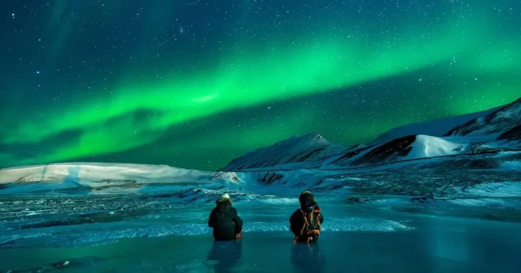 Two people stand in awe, gazing at Alaska’s northern lights as vibrant green and purple hues dance across the night sky—capturing the best time to visit for this breathtaking phenomenon.