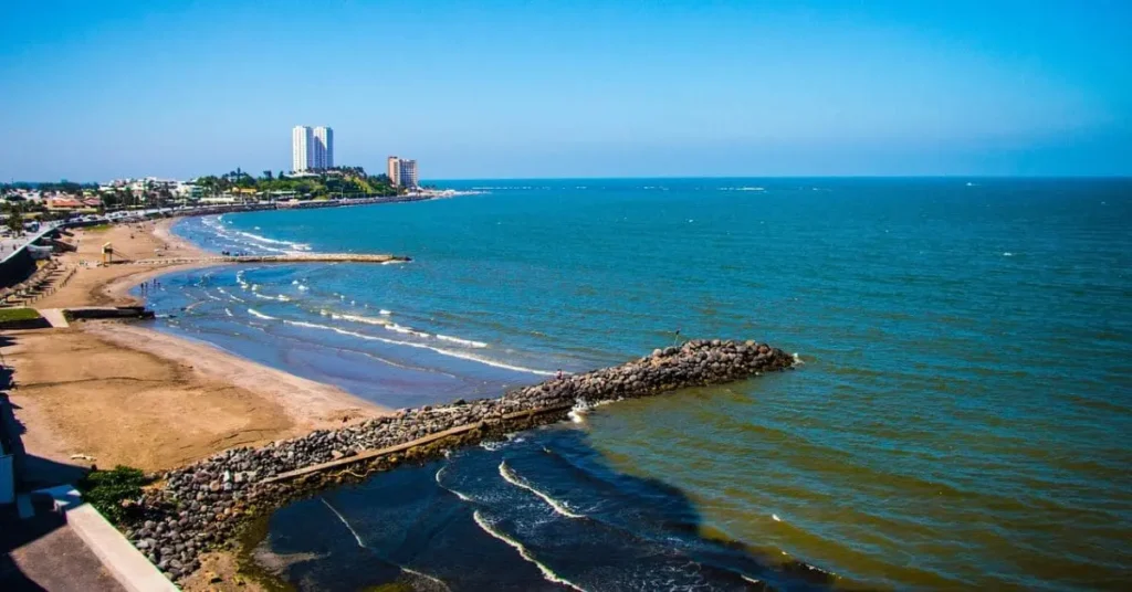 A panoramic view of the coastline in Veracruz, Mexico, showcasing a sandy beach, deep blue ocean, and a cityscape with tall buildings, suggesting the need to consider when is the best time to visit Veracruz for optimal weather and experience.