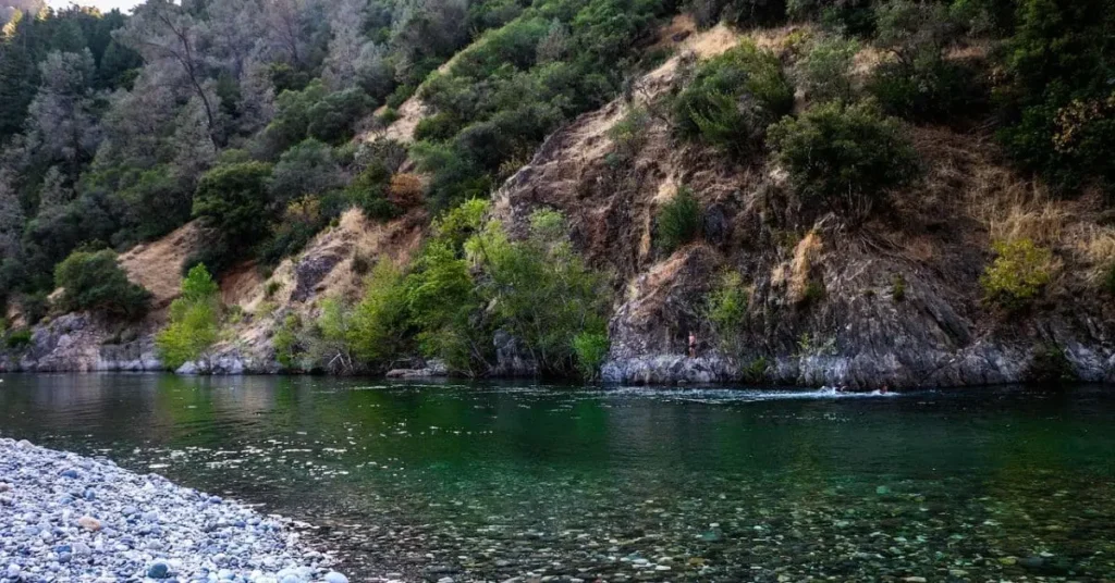 A serene view of Lake Clementine with a rocky shoreline, clear green water, and a forested hillside, showcasing the natural beauty of the area.