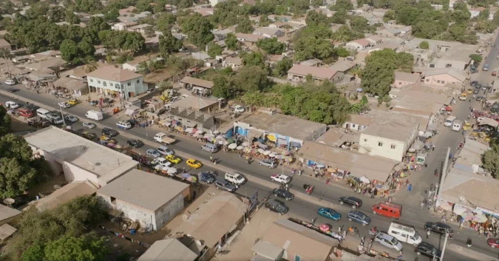 Aerial view of a bustling street in Senegal, showcasing local markets, vehicles, and residential buildings surrounded by lush greenery.