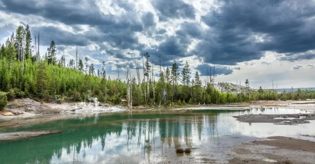 A scenic view of a hot spring surrounded by lush greenery and a dramatic cloudy sky in Yellowstone National Park, illustrating the beauty of the park for those researching where to stay when visiting Yellowstone.